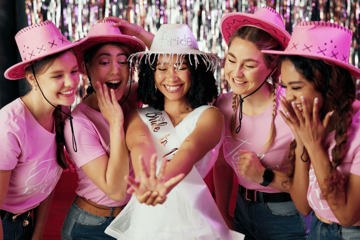 Group of friends at a bachelorette party in pink shirts and hats, center woman in a bridal sash laughing and high-fiving her friends.