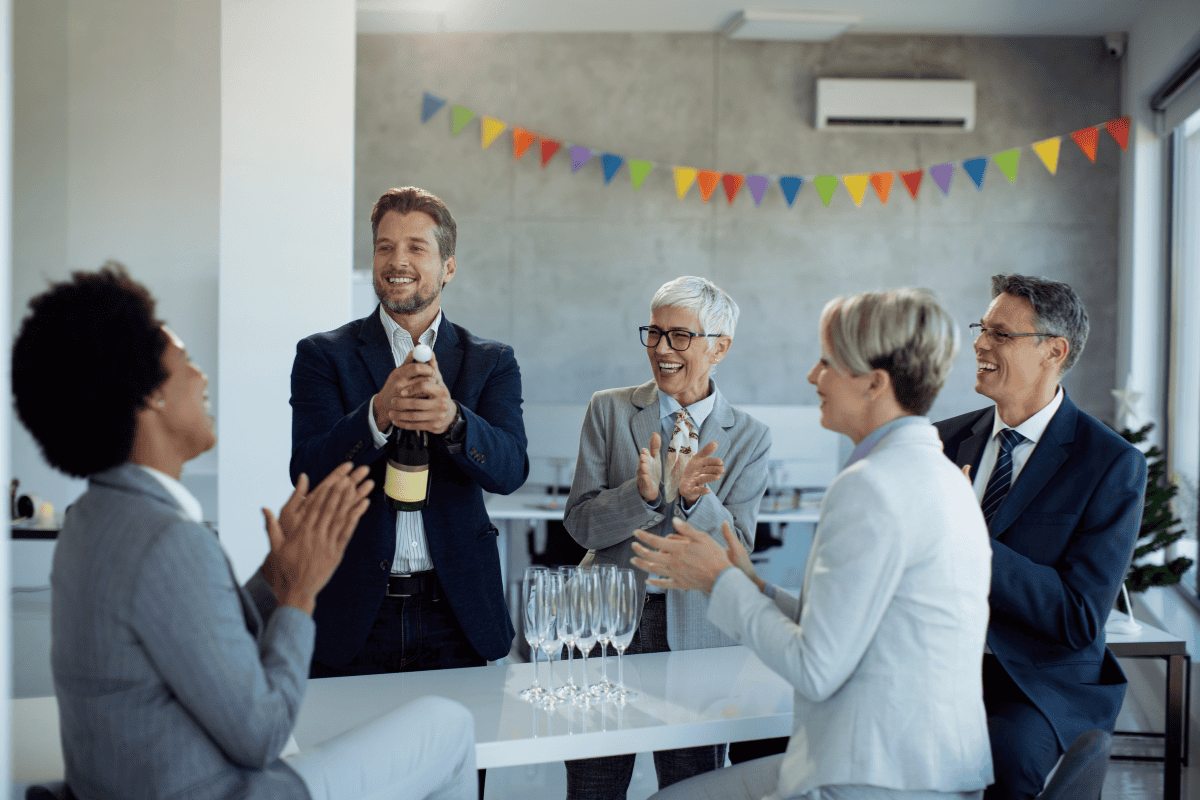 A group of professionals in office attire celebrate, clapping as a man opens a champagne bottle at a high-top table with glasses, colorful bunting above.